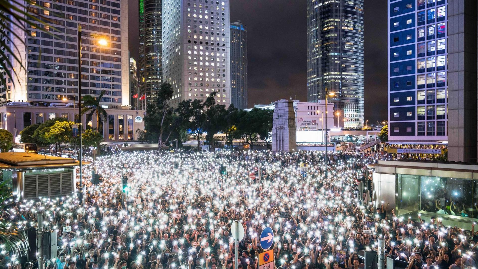 Nächtliche Demonstrantenversammlung mit Smartphones als Taschenlampen, während einer Kundgebung im Chater Garden, am 2. August 2019 in Hong Kong. Im Hintergrund sind beleuchtete Wolkenkratzer zu sehen.