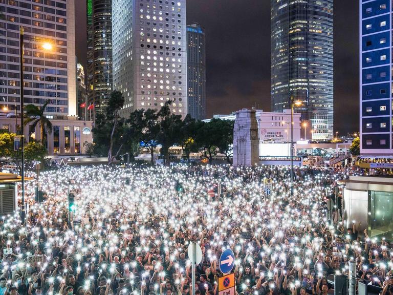 Nächtliche Demonstrantenversammlung mit Smartphones als Taschenlampen, während einer Kundgebung im Chater Garden, am 2. August 2019 in Hong Kong. Im Hintergrund sind beleuchtete Wolkenkratzer zu sehen.