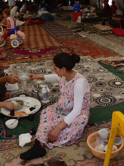 Eine yezidische Familie hat Schutz gefunden in einem Flüchtlingscamp in der Nähe der türkisch-irakischen Grenze. Erwachsene und Kinder sitzen auf ausgebreiteten Teppichen in einem Zelttunnel.