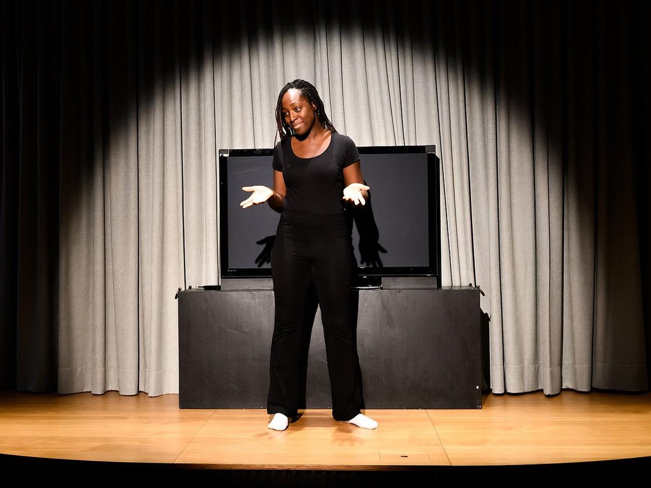Sarah Quarshie, eine Schwarze Frau, steht barfuß im Scheinwerferlicht eine Bühne. Sie hält fragen ihre Hände vor den Körper und ist einheitlich in schwarz gekleidet. 