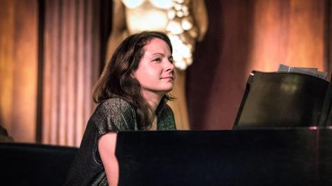 Vor einem schwarzen Piano sitzt eine dunkelhaarige junge Frau. Ihr Blick schweift zur Seite und im Hintergrund ist verschwommen eine Statue zu sehen.