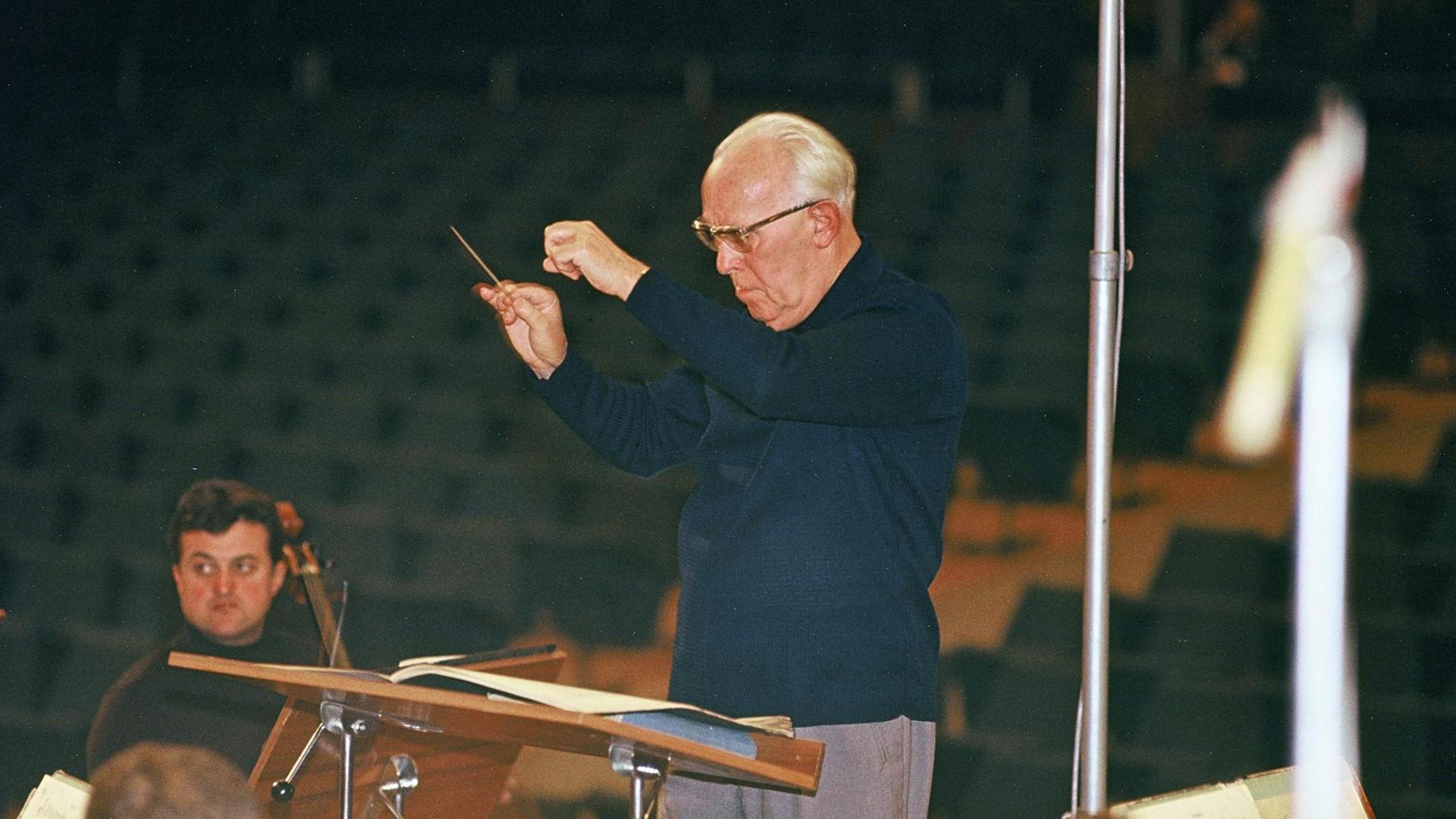 Der Dirigent Heinz Bongartz dirigiert eine Probe, er trägt eine helle Hose und einen blauen Rollkragenpullover, in der rechten Hand hält er einen kleinen Taktstock