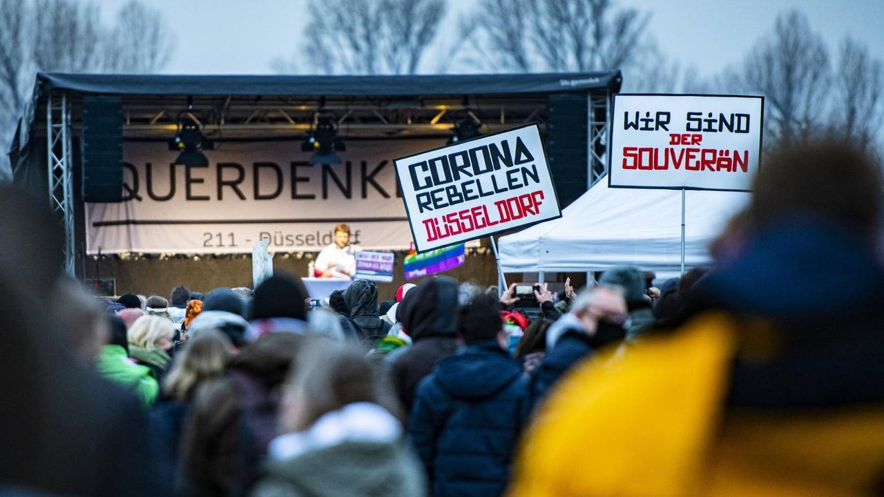 Eine Menschenmenge steht vor einer Bühne. Einige tragen Plakate, etwa mit der Botschaft: "Corona Rebellen Düsseldorf", und "Wir sind der Souverän".