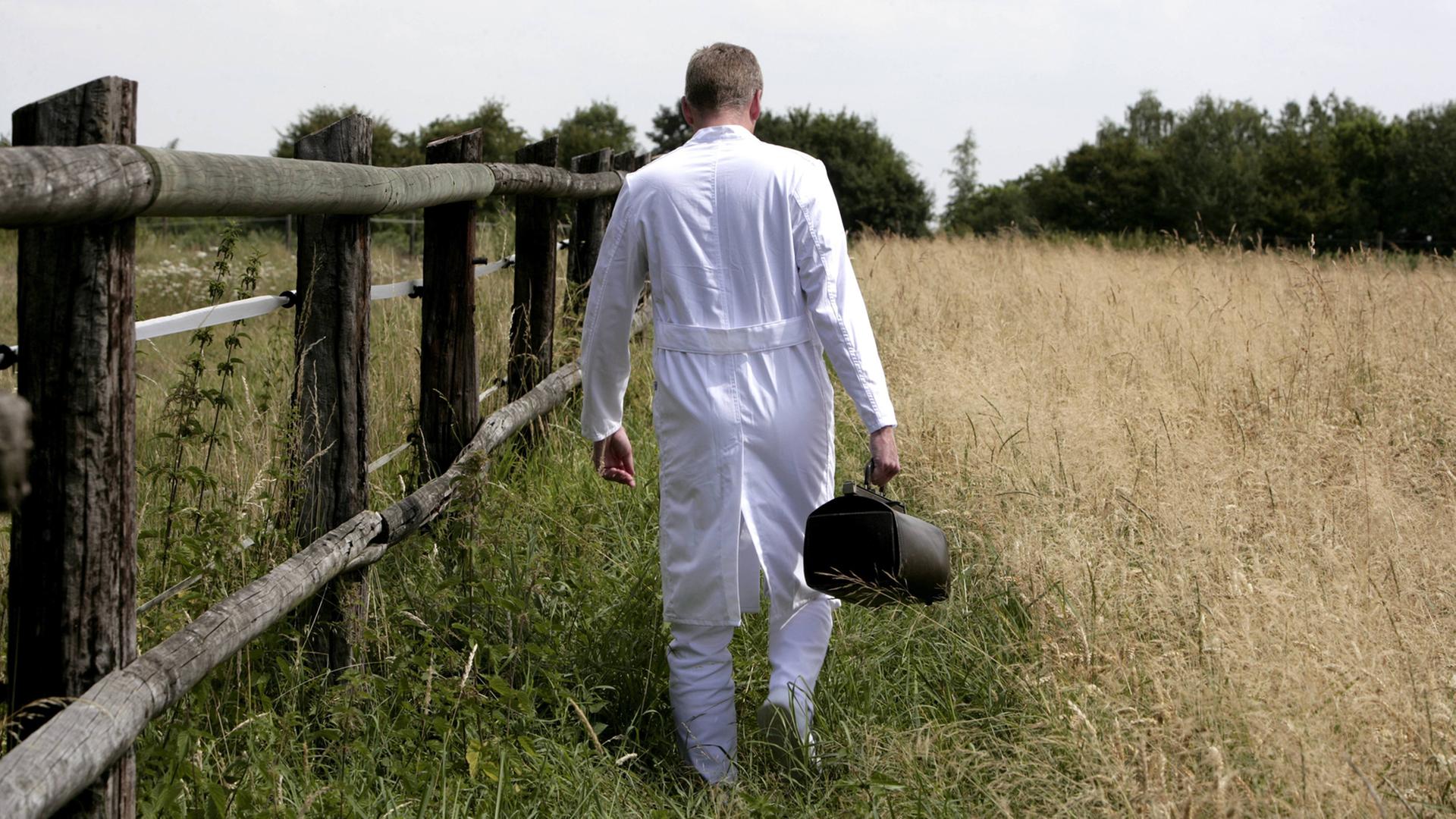 Ein Landarzt im weißen Kittel mit Ledertasche geht zwischen einem Pferdegatter und einem Kornfeld.