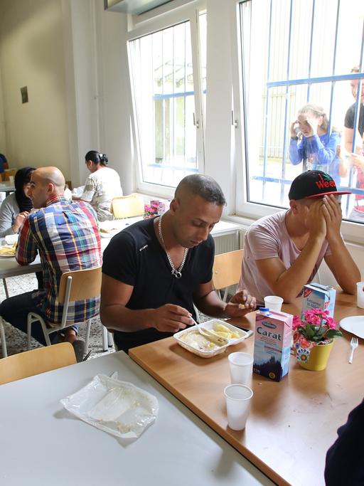 Flüchtlinge sitzen in einem Speisesaal an langen Tischreihen beim Essen zusammen.