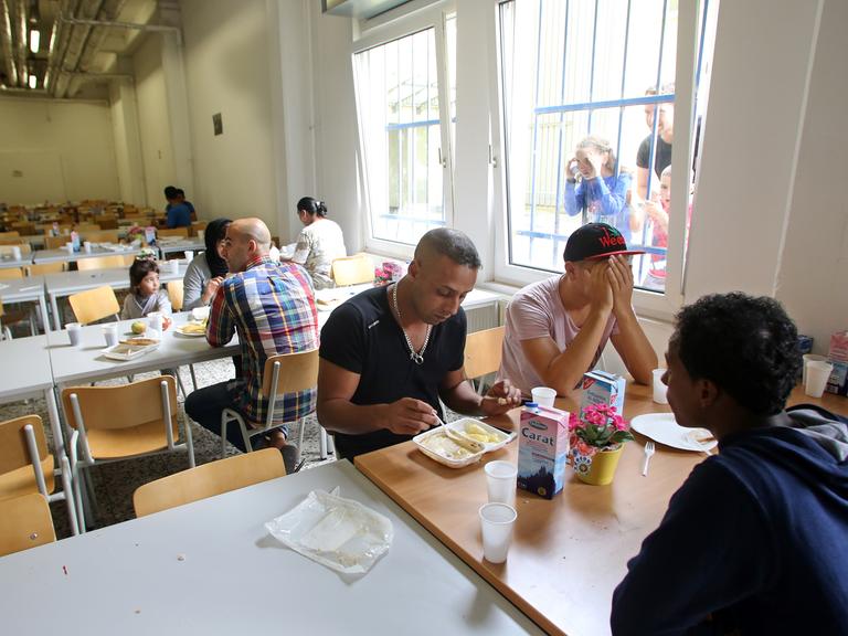 Flüchtlinge sitzen in einem Speisesaal an langen Tischreihen beim Essen zusammen.
