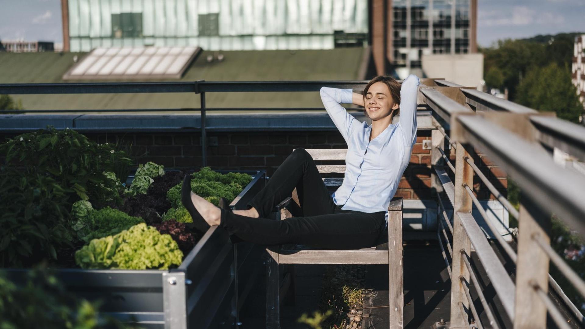 Eine Frau in Hemd und Geschäftshose hat auf einem Dachgarten die Beine hochgelegt und entspannt in der Sonne.