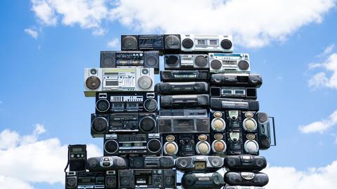 Aufeinander gestapelte Radios mit einem blauen Himmel und weißen Wolken im Hintergrund.