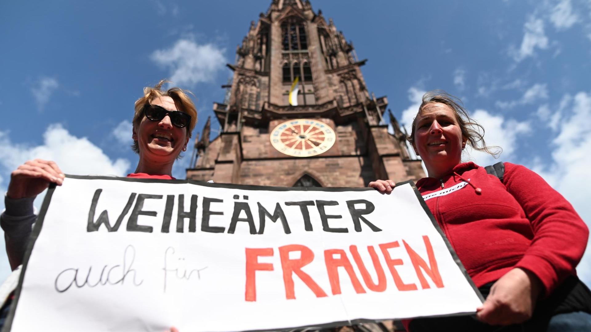 Zwei Frauen der Initiative Maria 2.0 der Katholische Frauengemeinschaft in der Erzdiözese Freiburg halten vor dem Freiburger Münster ein Banner mit der Aufschrift "Weiheämter auch für Frauen" in die Höhe.
