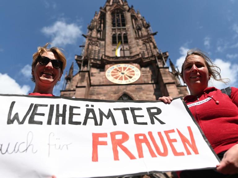 Zwei Frauen der Initiative Maria 2.0 der Katholische Frauengemeinschaft in der Erzdiözese Freiburg halten vor dem Freiburger Münster ein Banner mit der Aufschrift "Weiheämter auch für Frauen" in die Höhe.