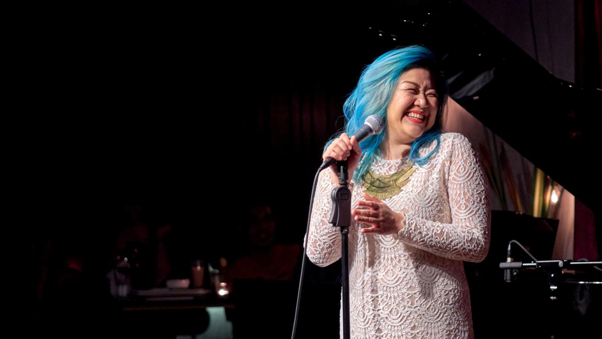 Die Sängerin Bihzhu, blau gefärbtes Haar, weißes Spitzenkleid, am Mikrofon auf der Bühne - sie schaut zur Seite und lacht