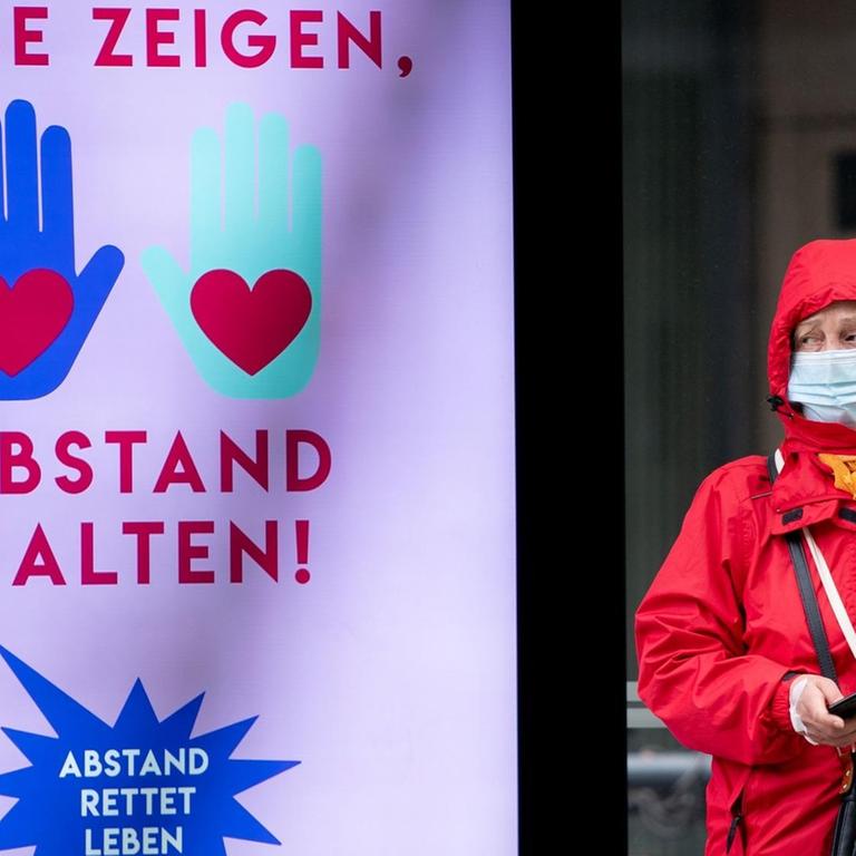 Eine Passantin steht mit einem Mund- und Nasenschutz an einer Straßenbahnhaltestelle neben einer Leuchtreklame mit der Aufschrift "Liebe zeigen, Abstand halten!"
