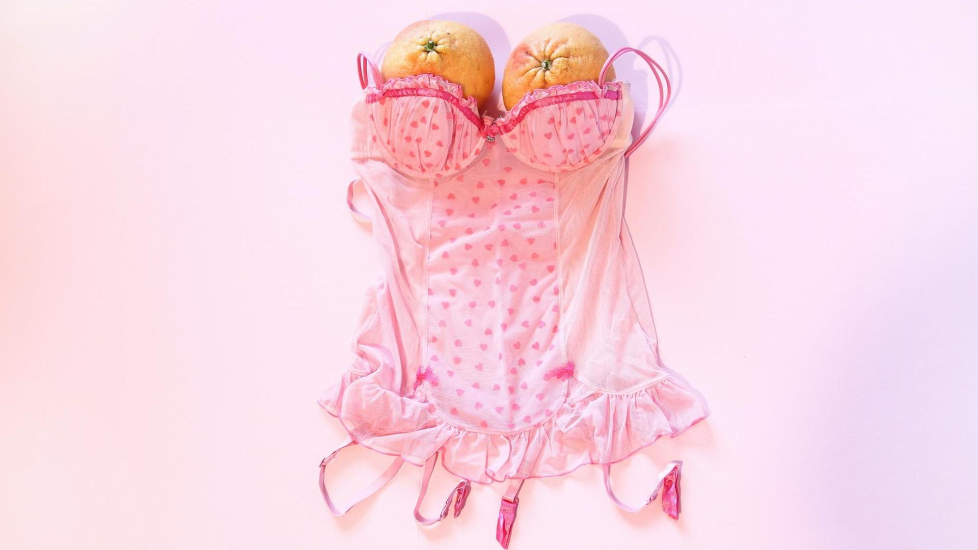 Eine rosa Korsage liegt auf rosa Hintergrund – dort wo beim Tragen die Brüste wären, sind zwei Orangen platziert.