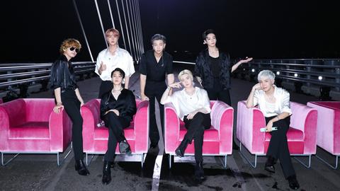 Die sieben Mitglieder von BTS auf einer Brücke. Einige sitzen in pinken Sesseln.