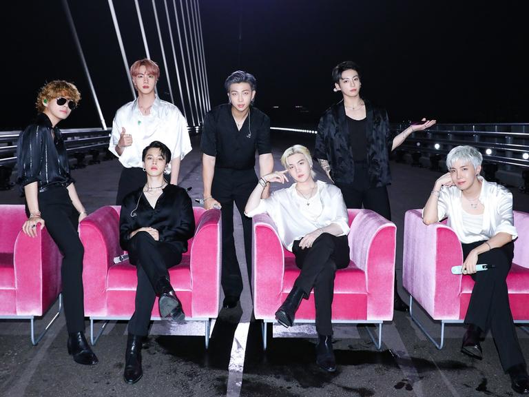 Die sieben Mitglieder von BTS auf einer Brücke. Einige sitzen in pinken Sesseln.