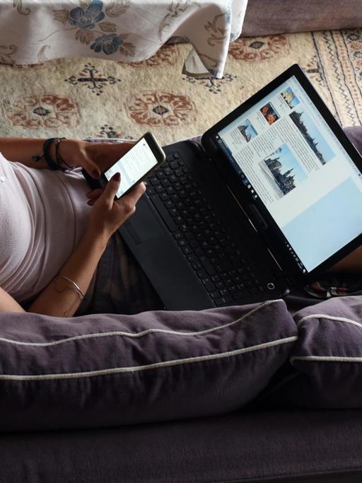 Ein Frau liegt auf einem Sofa und surft mit einem Smartphone und einem Laptop im Internet.
