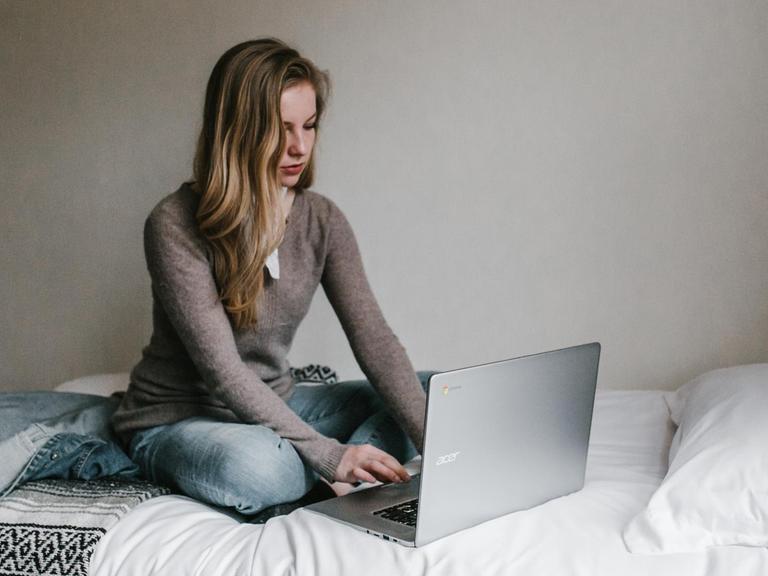 Eine junge Frau sitzt mit ihrem Laptop auf dem Bett.