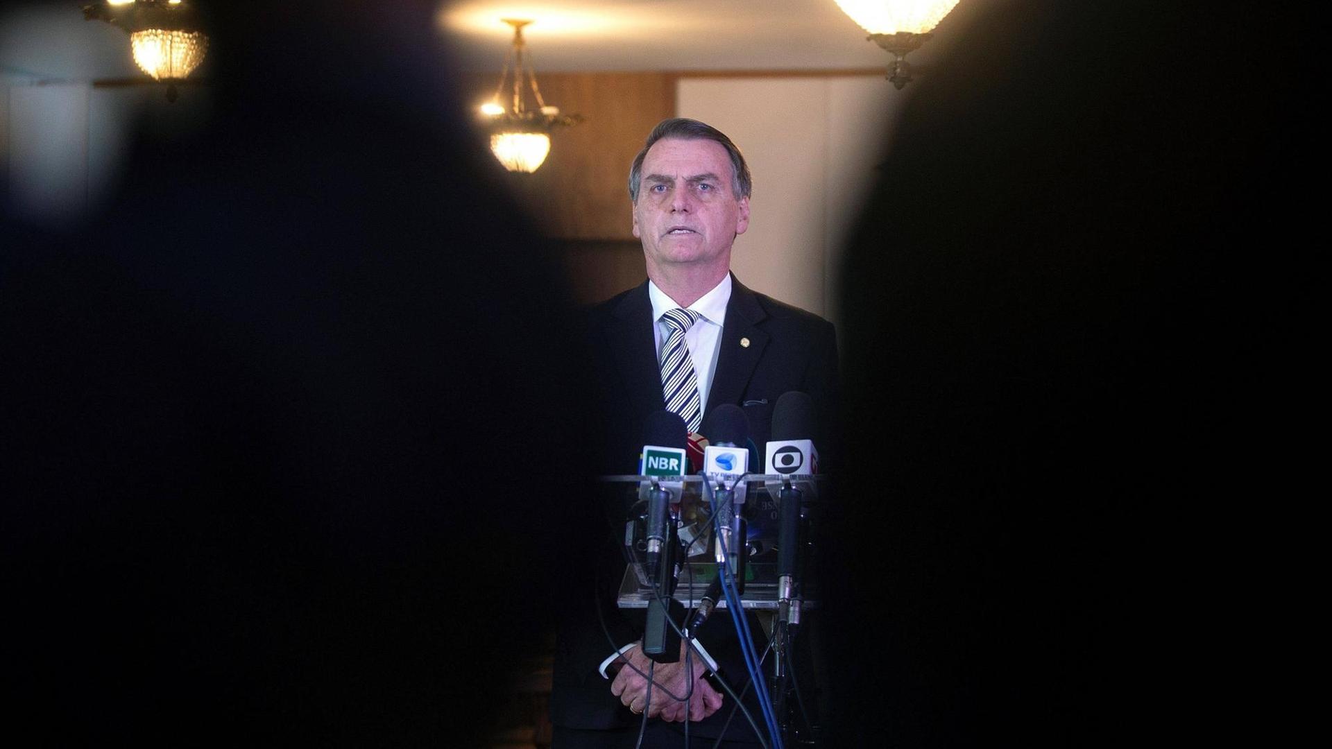 Durch eine schmale Lücke zwischen zwei Personen im Vordergrund ist Brasiliens Präsident Jair Bolsonaro zu sehen. Vor ihm stehen mehrere Mikrofone.