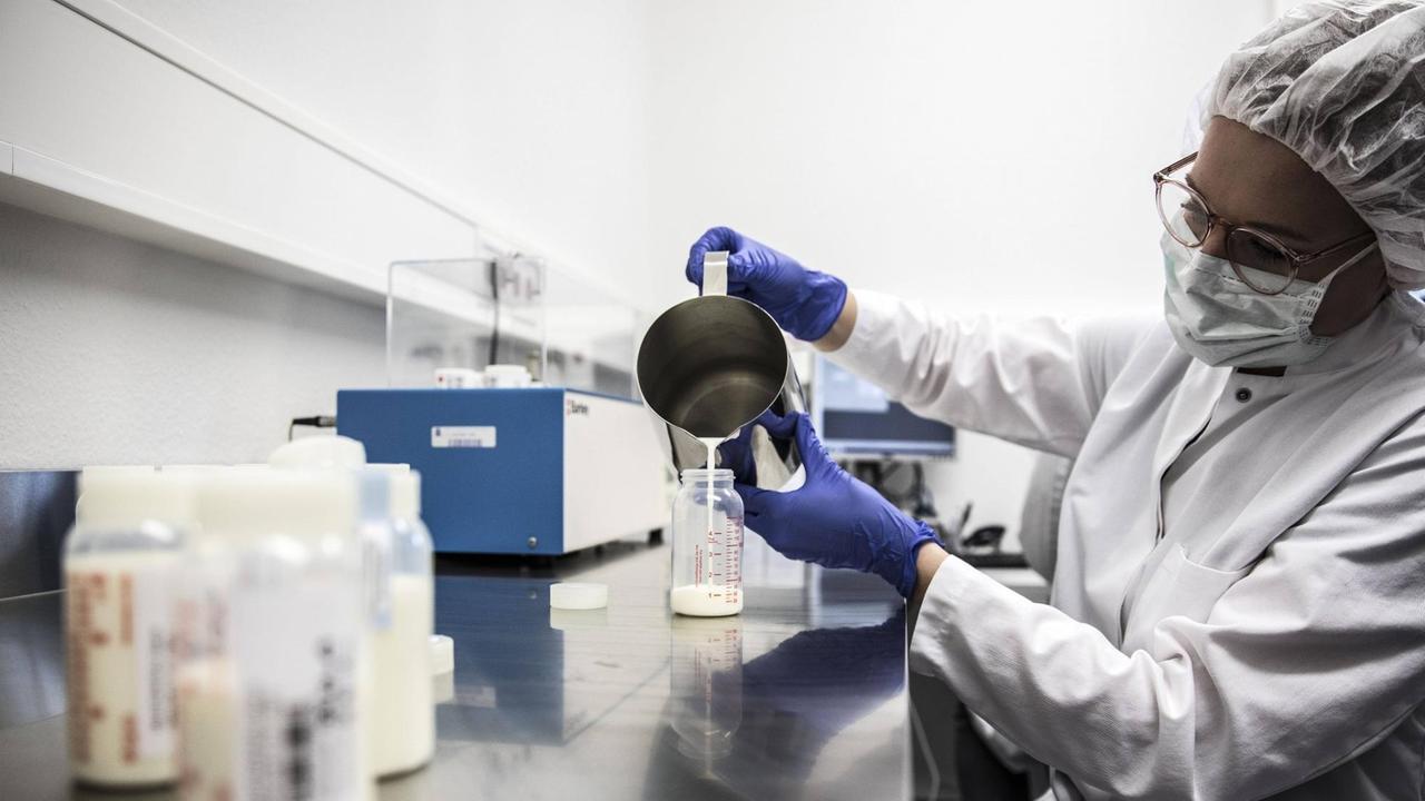 Eine Labormitarbeiterin gießt Milch aus einem Kännchen in eine schmale Glasflasche.