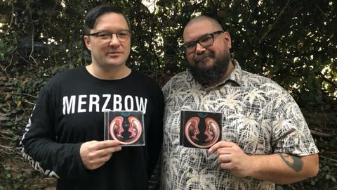 Auf dem Bild sind die Macher des Merzcasts Mike Connelly und Greh Holger zu sehen. Sie halten eine CD von Merzbow in der Hand und lächeln in die Kamera