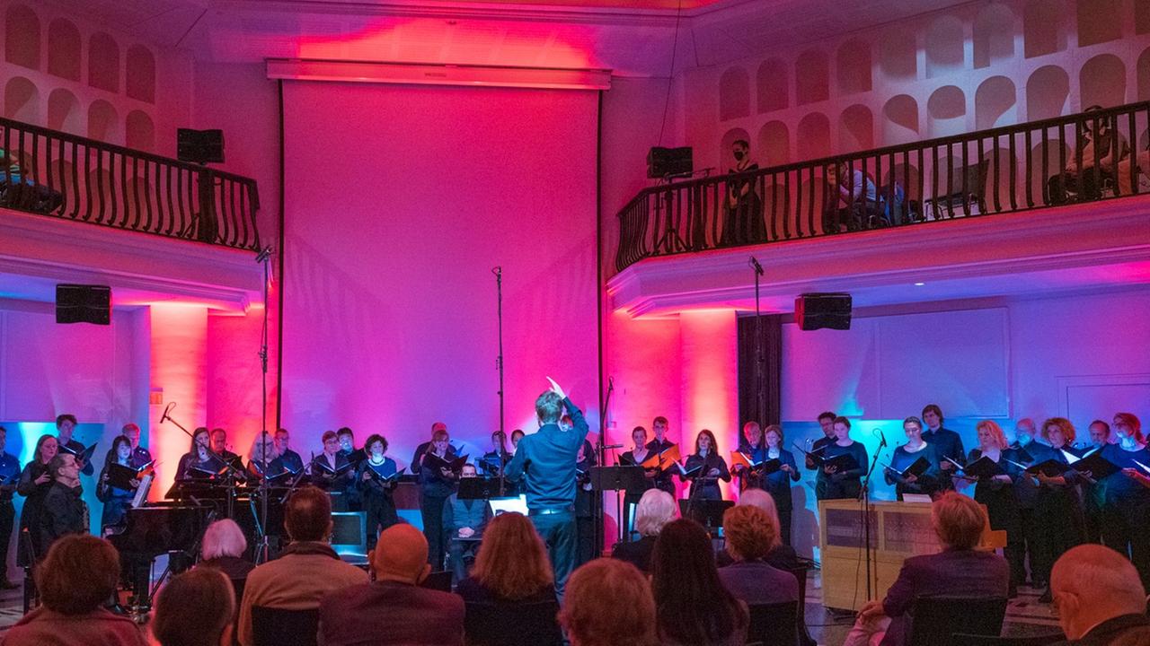 Das Publikum sitzt verstreut vor dem aufgestellten Rundfunkchor Berlin im lila-pink ausgeleuchteten silent green.