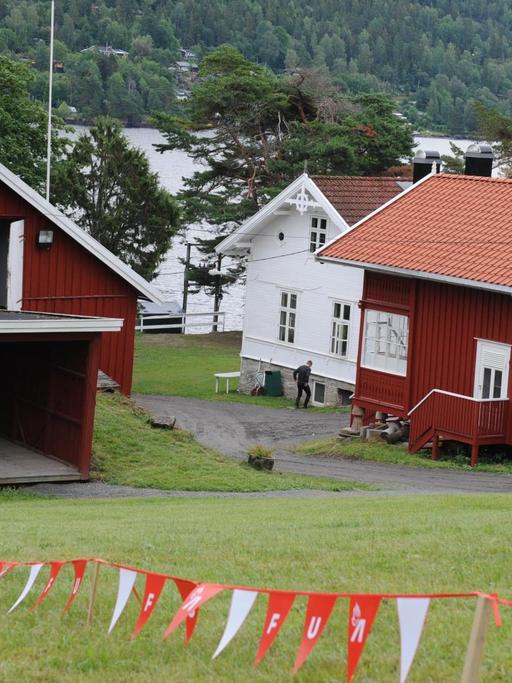 Drei Gebäude auf der norwegischen Insel Utøya. Im Vordergrund flattert ein rot-weißes Absperrband mit den Buchstaben AUF. Sie stehen für die Jugendorganisation der norwegischen Arbeiterpartei. Im Hintergrund sind Wasser und Bäume zu sehen.