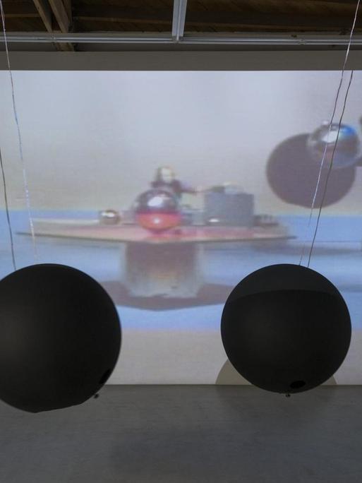 Große Bälle pendeln auf einer Bühne, im Hintergrund einer Videoprojektion mit drei Spielern und den Ballschatten