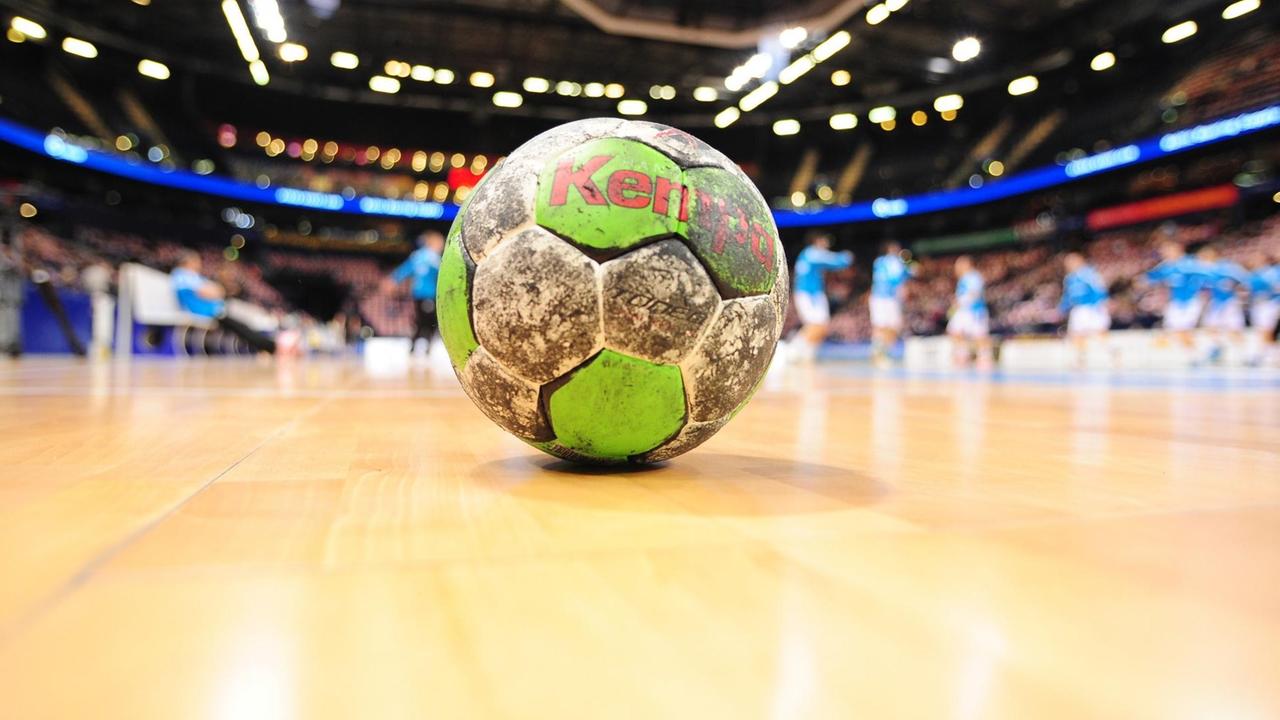 Das Bild zeigt einen schmutzigen grün-weißen Handball, der auf dem Parkett des Platzes liegt. Im Hintergrund unscharf Spieler und die Zuschauerränge.