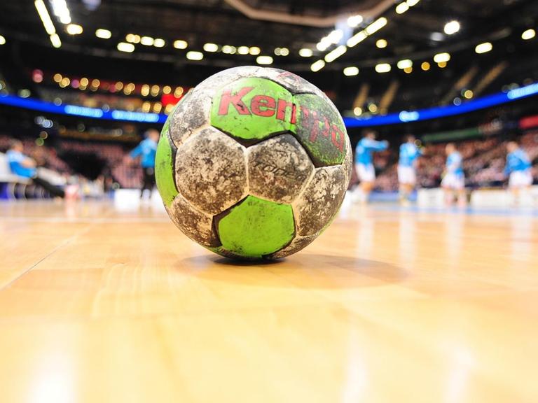 Das Bild zeigt einen schmutzigen grün-weißen Handball, der auf dem Parkett des Platzes liegt. Im Hintergrund unscharf Spieler und die Zuschauerränge.