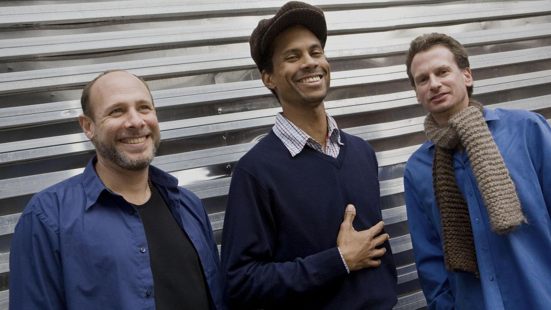Drei Männer stehen lachend nebeneinander vor einem Garagentor. Der rechte und linke tragen jeweils ein blaues Hemd, der mittlere eine große Ballonmütze