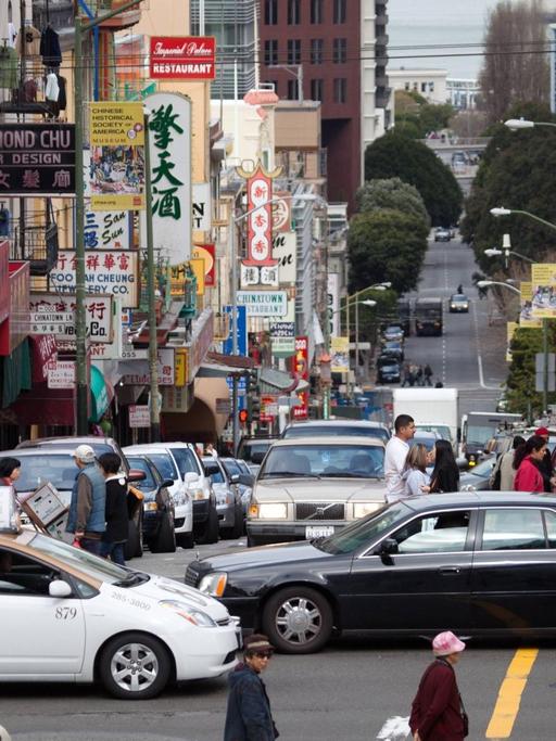 Kreuzung in Chinatown in San Francisco, Menschen und Autos auf der Straße, an den Gebäuden Schilder mit chinesischer Schrift.