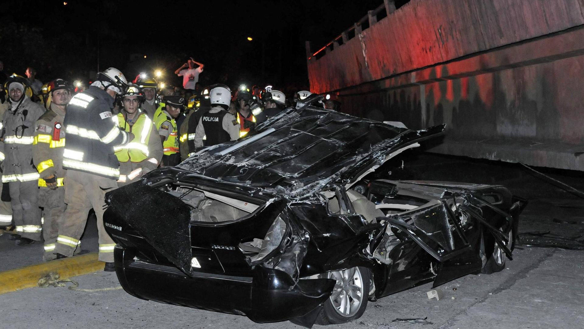 Ein Auto wird von einer zusammengebrochenen Brücke zerstört, Rettungskräfte im Hintergrund.