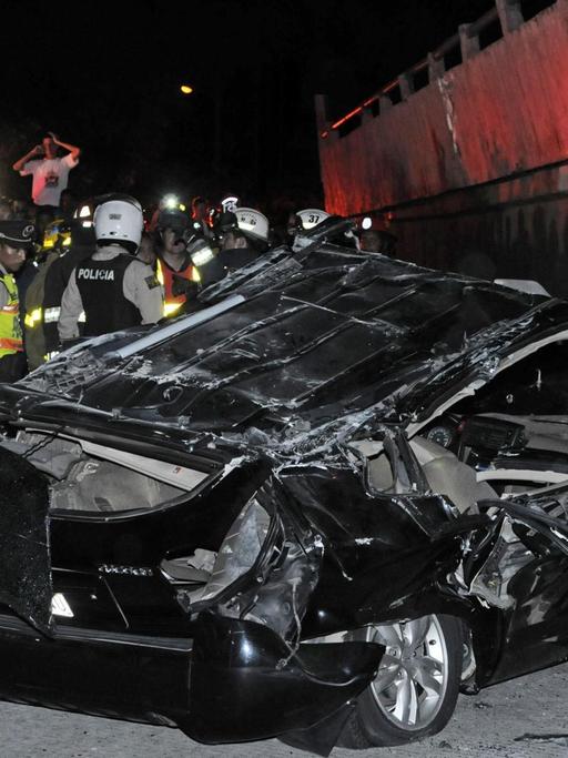 Ein Auto wird von einer zusammengebrochenen Brücke zerstört, Rettungskräfte im Hintergrund.