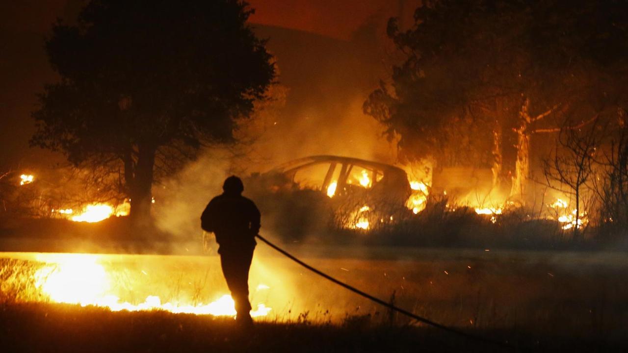 Helle Flammen schlagen aus einem Wald. Darin steht ein verbranntes Auto. Ein Feuerwehr-Mann löscht. 