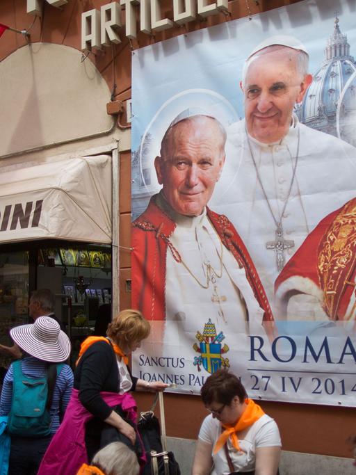 Menschen stehen vor einem Plakat mit den Päpsten Franziskus, Johannes XXIII. und Johannes Paul II.