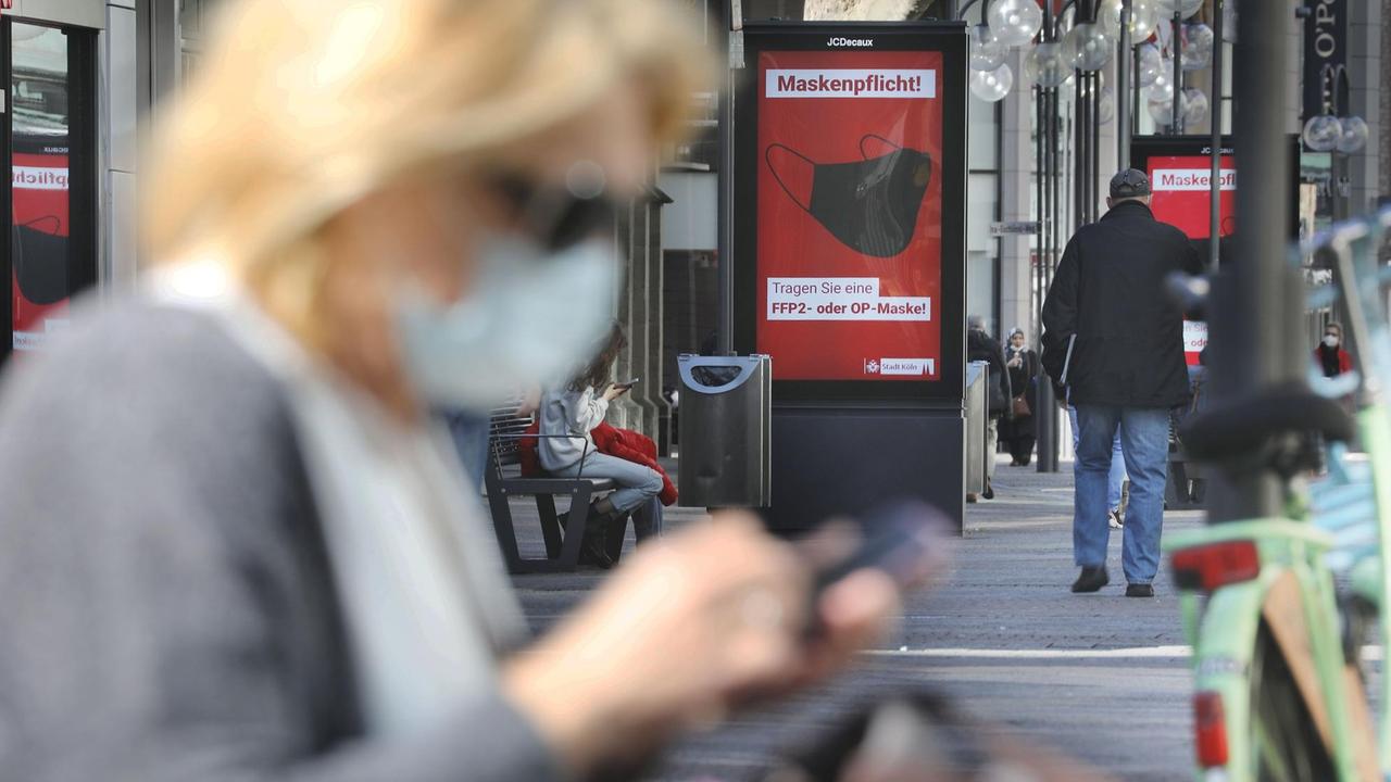 Eine einsam wirkende Frau sitzt mit Maske in der Fußgängerzone vor einer Anzeigetafel, die auf die Maskenpflicht hinweist.