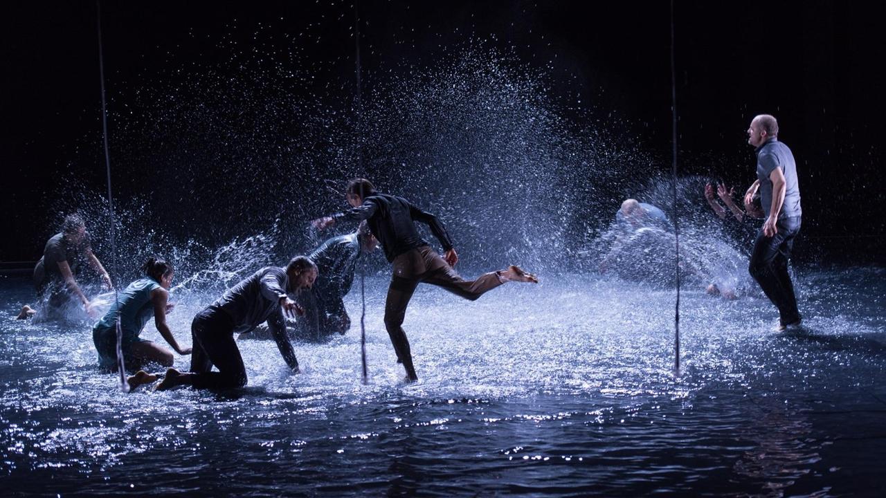 Mut und Gnade Regie: Luk Perceval Ensemble Foto: Robert Schittko
Ensemblemitglieder in einer dynamischen Szene mit vom Boden spritzendem Wasser im Halbdunkel auf der Bühne.