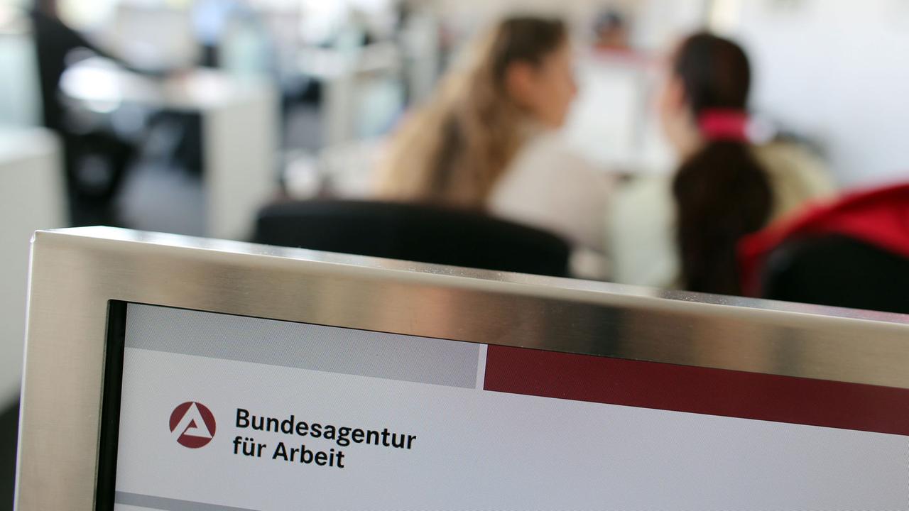 Blick in ein Jobcenter: im Vordergrund das Logo der Bundesagentur für Arbeit, im Hintergrund verschwommen zwei Frauen, die miteinander reden