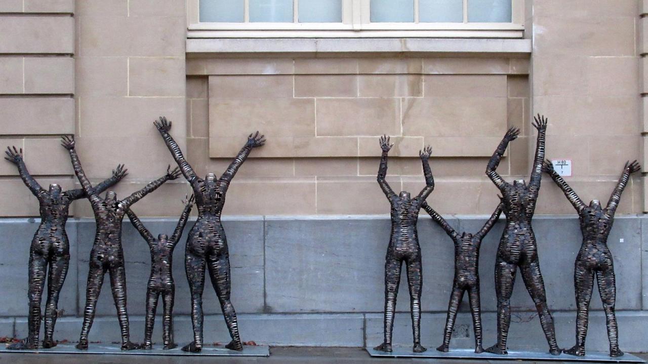 Graue Skulptur recken die Arme nach oben und stehen an einer Außenwand des Museums.