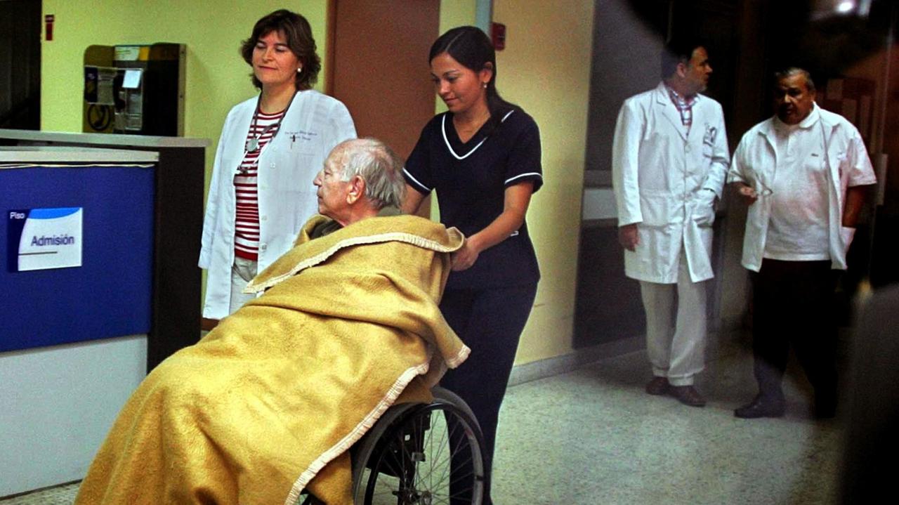 In einem Rollstuhl unter einer gelben Wolldecke sitzend wird Paul Schäfer durch das Polizeikrankenhaus in Santiago gefahren.