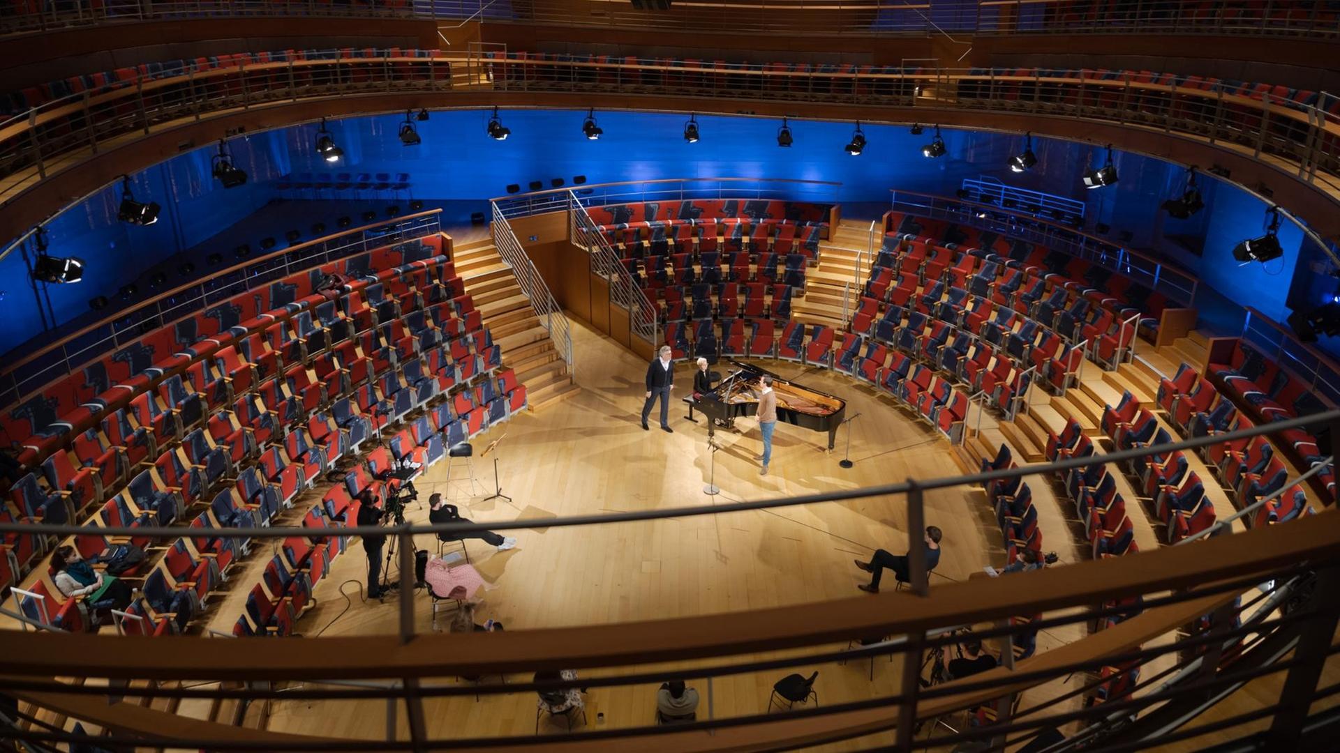 Saalansicht eines geschwungenen Konzertraumes mit roten Sesseln, die ovalförmig um die Bühne angeordnet sind. Auf dem Parkett steht ein Flügel, mehrere Menschen und zwei Kameras.