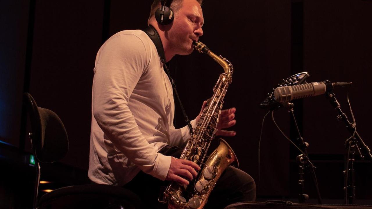 Ein junger Mann mit weißem Oberteil sitzt unter Kopfhörern vor einem Mikrofon und spielt Saxofon.