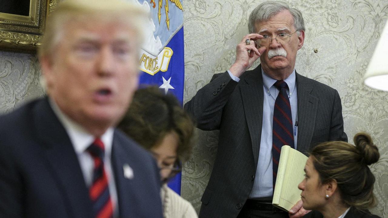 Man sieht Trump unscharf im Vordergrund in einem Raum, er sagt gerade etwas. Dahinter steht Bolton an einer Wand mit Dokumenten im Arm. Er rückt sich die Brille zurecht. Ihn sieht man scharf. Man sieht Trump unscharf im Vordergrund in einem Raum, er sagt gerade etwas. Dahinter steht Bolton an einer Wand mit Dokumenten im Arm. Er rückt sich die Brille zurecht. Ihn sieht man scharf.