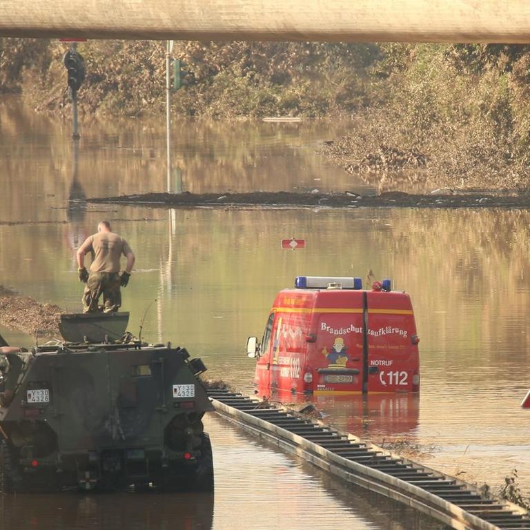 Blick auf die überflutete B265 bei Erftstadt Liblar. Ein Panzer der Bundeswehr zieht einem Wagen aus der Flut. Ein Einsatzwagen der Feuerwehr und ein LKW stehen noch unter Wasser.