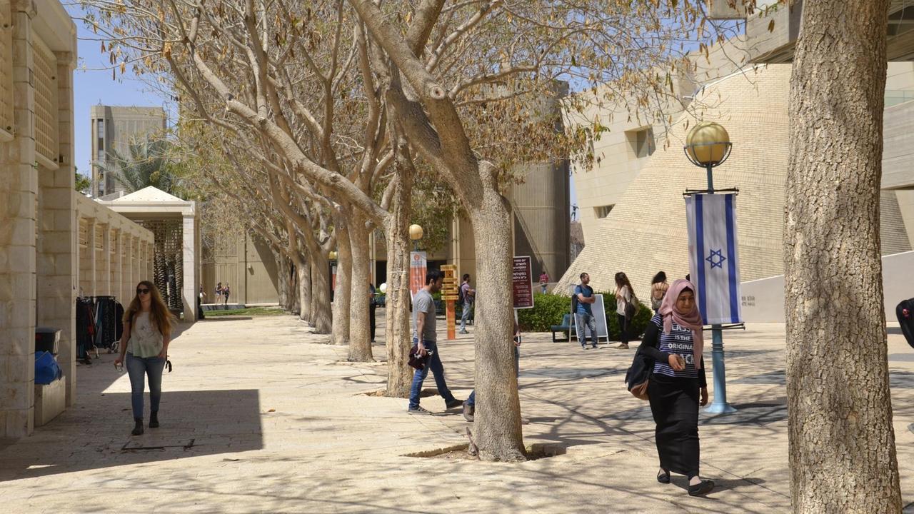Mehrere Studenten mit Büchern und Rucksäcken laufen über den Campus der Universität. Es stehen Bäume auf dem Weg.
