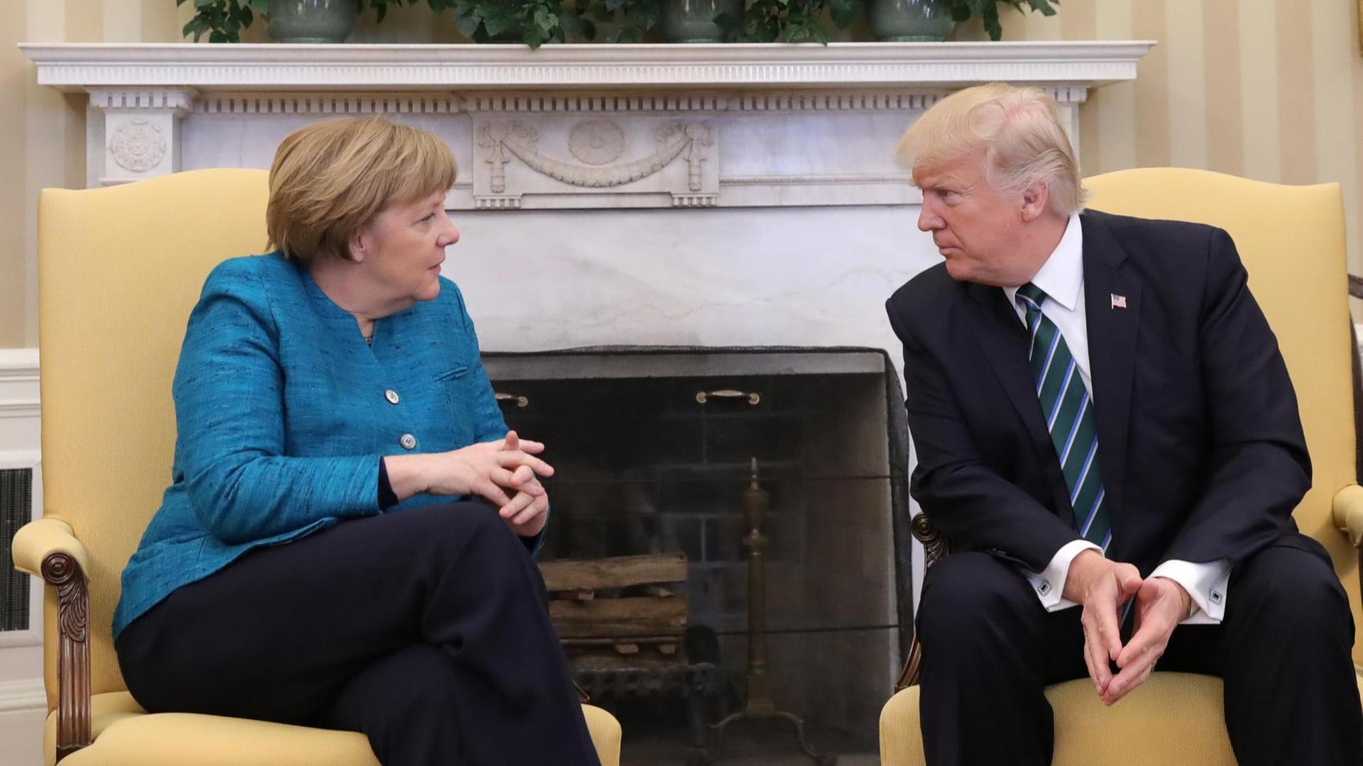 Sie sehen die Bundes-Kanzlerin und den Präsidenten von dem Land USA. Sie sitzen vor einem Kamin.