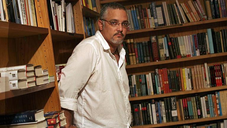 Der kubanische Schriftsteller Enrique del Risco steht vor einer Bücherwand und schaut in die Kamera.