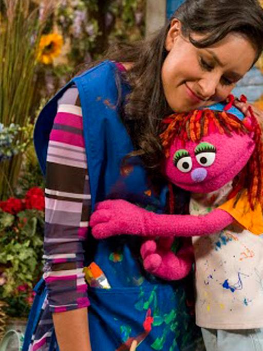 Die Puppe Lily aus der Fernsehserie Sesamstraße, die mit ihrer Familie obdachlos wird, schmiegt sich an eine Frau.