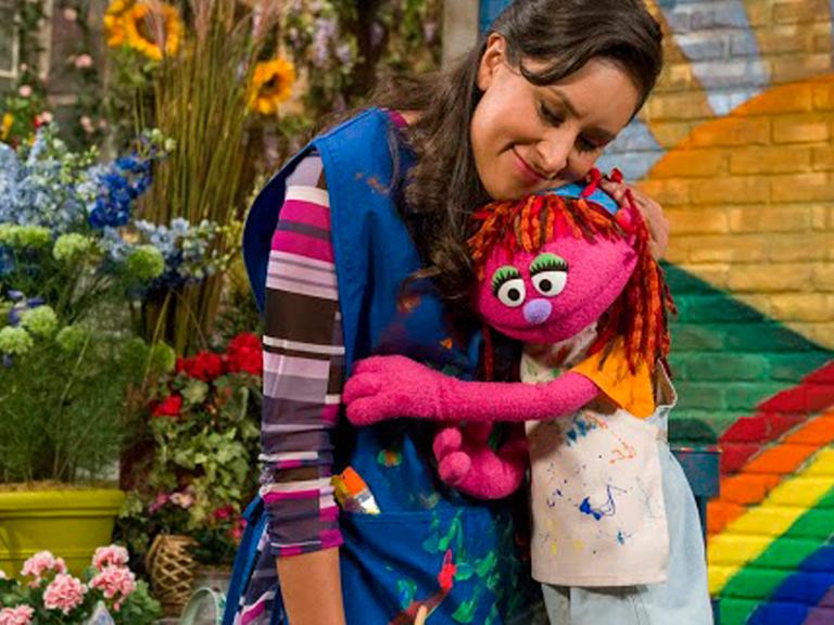Die Puppe Lily aus der Fernsehserie Sesamstraße, die mit ihrer Familie obdachlos wird, schmiegt sich an eine Frau.