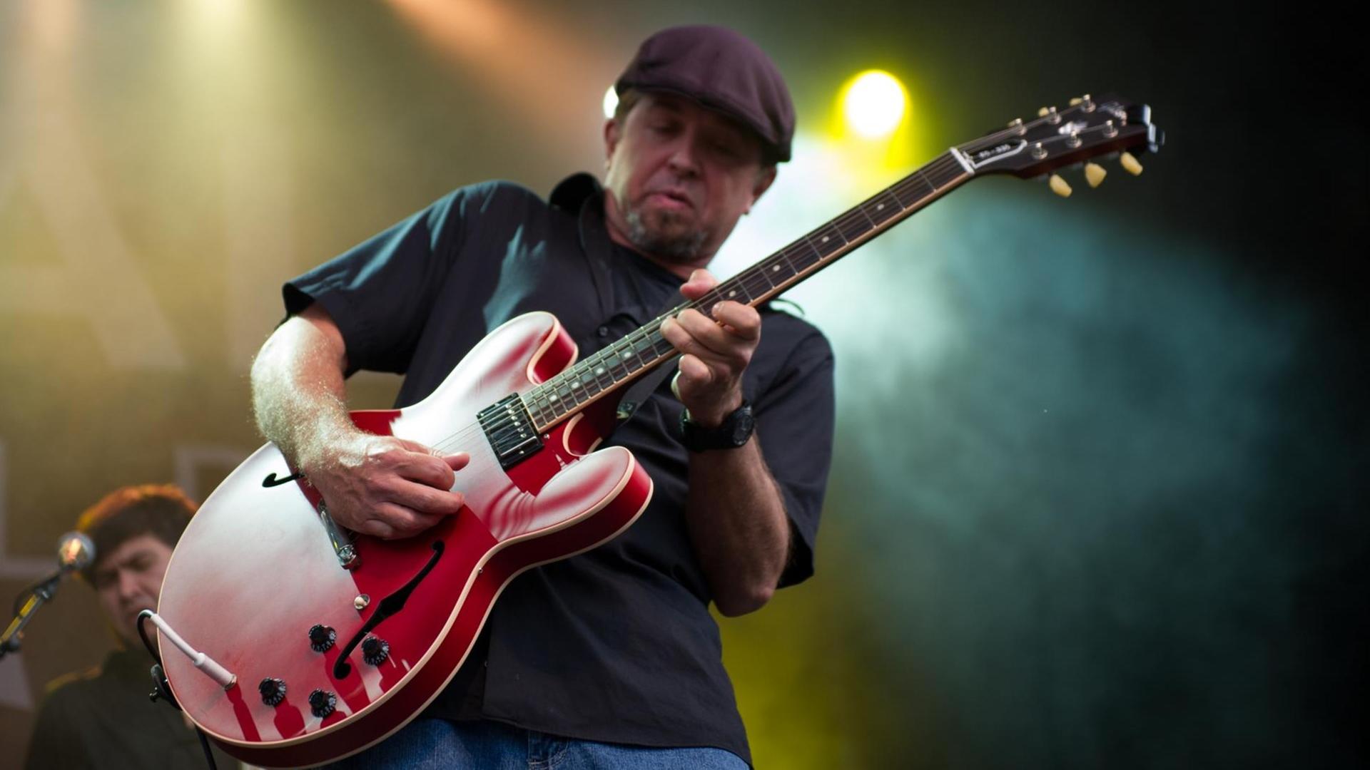 Ein Mann mit Kappe steht auf einer Bühne und spielt eine rote Gitarre.